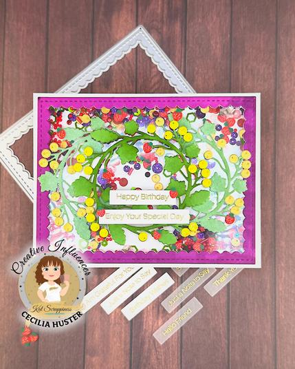 A handmade birthday card with a vibrant purple border featuring a decorative design of colorful paper fruits including strawberries and citrus fruits arranged in a wreath pattern with green leaves. The card displays "Happy Birthday" and "Enjoy Your Special Day" messages in the center. 

The card is photographed on a wooden surface alongside craft supplies including sentiment strips with phrases like "Happy Birthday," "Thinking of You," "Just a Note to Say," "Thank You," and "Hello Friend." In the bottom left corner is a circular logo for "Creative Influencer Cecilia Huster by Kat Scrappiness." 

Small decorative strawberry embellishments are scattered around the card.