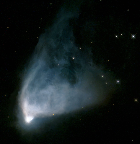 A Hubble Space Telescope image of a fan-shaped reflection nebula. At the lower left, a bright white star illuminates a surrounding cloud of blue and gray gas and dust, which spreads outward in a triangular formation. The nebula fades into darkness toward the upper right, surrounded by a scattering of stars on a black background.