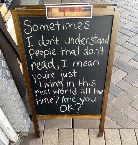 Sandwich blackboard sign on the paved footpath outside Q's Books at Hamilton with chalk writing: "Sometimes I don't understand people that don't read, I mean you're just Living in this real world all the time? Are you OK?