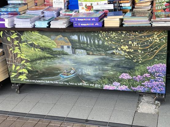 There is a mural across the lower front of a book display table, maybe 2m or so across and 1m high. Ratty is rowing a happy Mole up the river in an idyllic English country scene with a cottage on the left bank and purple flowers to the right.  

White painted text quote from Wind in the Willows "It was so very beautiful that the mole could only hold up both paws and gasp. Oh my! oh my! oh my!" Artist is only given as Flying Spanners Gallery, Teralba.