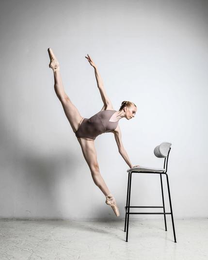Ballerina Polina Kokhan does a split jump with her weight pushed with one hand on a chair. The photo was taken at the Pavilion Studio St Petersburg, using a white background as a good contrast to the muscle structure of her arms and legs and the coffee coloured leotard she wears.
