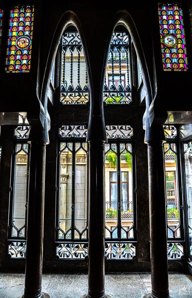 View looking out through tall parabolic arches atop columns and modernisme styled windows, some with stained glass, behind them. Through the windows the facade of the building across the street can be seen.