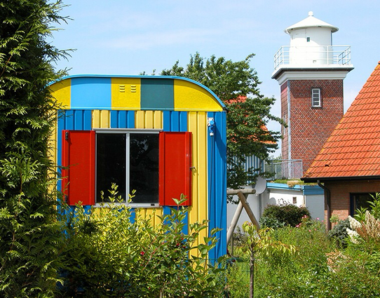 Ein gelb-blau gestichener Bauwagen mit roten Fensterläden. Rechts im Bild ein kleiner Leuchtturm und das Dach eines Hauses. Im Vordergrund und ganz links Pflanzen. In Schleswig-Holstein vor einigen Jahren.