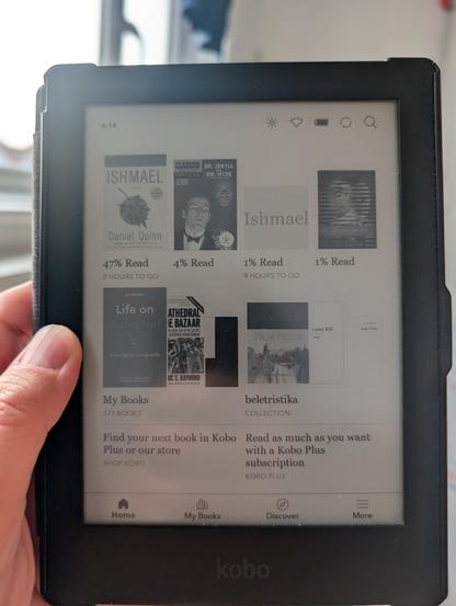 A close-up photo of a black Kobo Aura HD e-reader held in a hand. The screen displays the home interface with several book covers. The most prominent book, in the top left, is "Ishmael" by Daniel Quinn, showing 47% read. Other books visible include "Life on Autopilot" and "Cathedral & Bazaar". The Kobo logo is visible at the bottom of the device.