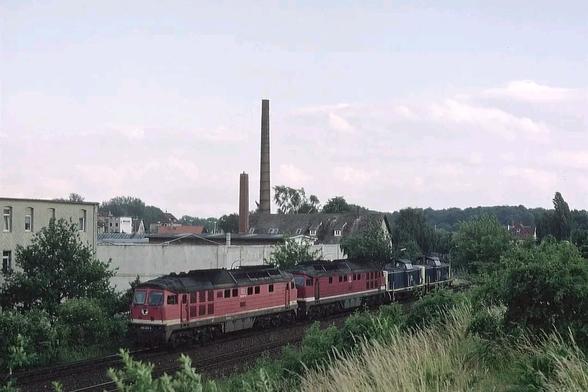 Ein Lokzug aus vier Diesellokomotiven durchquert eine gemischte Stadtrandlandschaft. Die beiden führenden Lokomotiven sind rot lackiert, gefolgt von zwei blau-beigen Maschinen. Links im Bild stehen ältere Wohn- und Fabrikgebäude, darüber ragen zwei schmale Industrieschornsteine auf. Der Zug fährt auf einem von Vegetation gesäumten Gleisbogen, im Vordergrund wie auch am rechten Bildrand wachsen dichte Sträucher und hohes Gras. Der Himmel ist leicht bewölkt, die Szenerie wirkt sommerlich ruhig und trägt den Charme vergangener Eisenbahnzeiten. Im Hintergrund zeichnen sich die Hügelzüge östlich von Braunschweig ab.