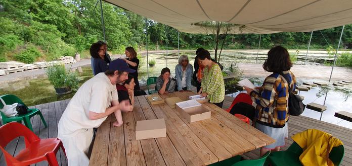 A group of people round a wooden table under a canopy with trees and water in the background