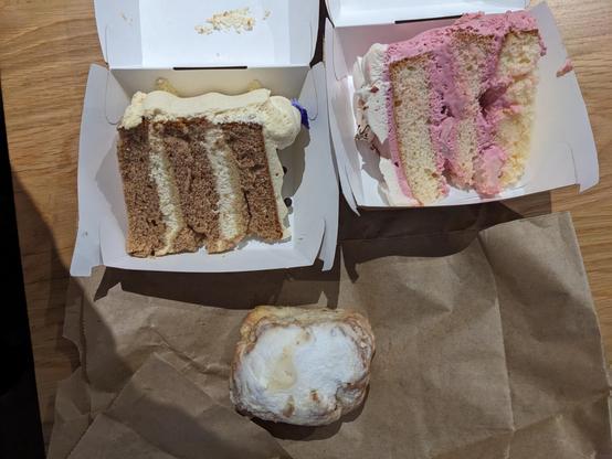 Aphoto of a tasty but at end of life scone, a wedge slice of heavily iced  carrot cake and a wedge slice of as yet undermined other cake, which is white cake with 3 layers of pink icing.