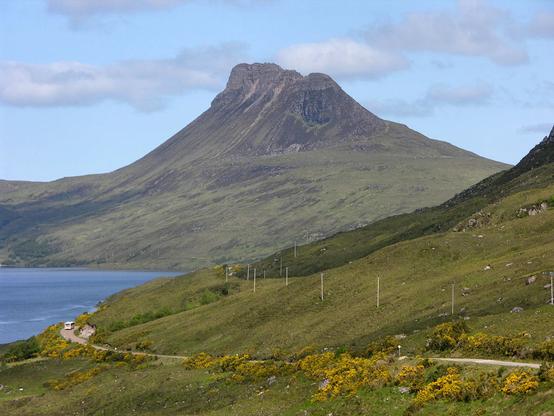 Stac Pollaidh. The image shows a mountain slope descending from right to left in the foreground with a narrow road leading around it. There’s a bus on the road. Further back and on the left is a body of water and in the distance is a mountain with a jagged summit ridge extending from right to left back into the frame. The scene is in sunlight.