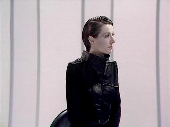 Claude 3.7 describes the image as: "This image appears to be from a science fiction television production, featuring a person in a sleek black uniform with a high collar and military-style design. The setting has a minimalist, futuristic aesthetic with vertical white panels forming the background, giving it a sterile, possibly spaceship or high-tech facility appearance. The subject is shown in profile, wearing their dark hair slicked back, which complements the austere, utilitarian style of their costume. The overall visual style suggests a dystopian or military science fiction setting, with clean lines and monochromatic color scheme typical of certain British sci-fi productions from the late 1970s to early 1980s."

Blakes7Bot, version 3.9.30.