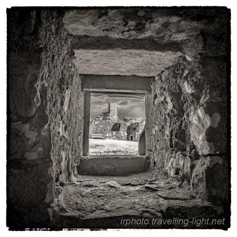 A toned black and white infrared photo looking out through a small square window in a thick stone wall to the courtyard of a ruined castle.