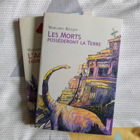 Les morts posséderont la Terre, une novella de Margaret Killjoy éditée dans la collection RéciFs chez Argyll. La couverture montre un bâtiment délabré devant lequel se tient un dinosaure