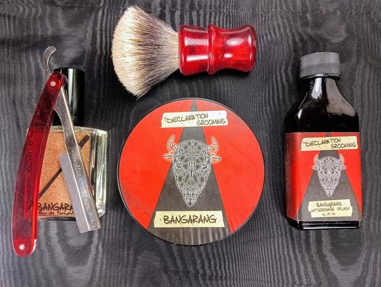 (Le Chat Mistral description)

The image showcases a collection of grooming items arranged neatly on a dark textured background. At the centre, there is a round tin labelled "Declaration Grooming" with a geometric bull's head and the word "Bangarang." To the left, there is a red-handled straight razor and a small bottle of cologne. Above the tin, there is a shaving brush with a red handle. To the right of the tin, there is a dark bottle of aftershave splash, also labelled "Declaration Grooming" and "Bangarang." The items are vibrant and well-organised, suggesting a traditional shaving setup.