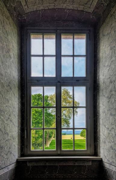 A rectangular, tall, wooden-framed window with 12 panes looks out onto a sunlit, verdant landscape. Through the upper panes, a bright blue sky with scattered white clouds is visible. The lower panes frame a view of lush green trees, with a large, light-green tree dominating the right side. Beyond the trees, a golden field stretches towards a distant blue horizon line. The window is set within a dark grey, subtly patterned wall, with a sill that matches the wall's texture. The overall impression is one of a serene, pastoral view from inside a historical building.
