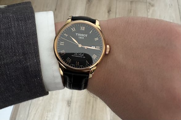 Tissot dress watch in black and gold, the wearer also wears a suit in grey and long sleeved shirt in white