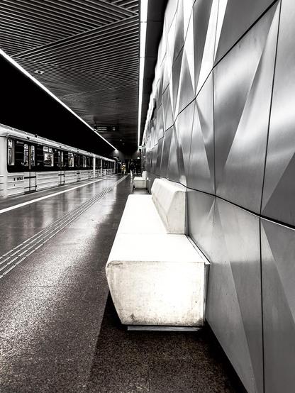 Ein schwarz-weißes Foto einer modernen U-Bahn-Station. Auf der linken Seite fährt ein langer Zug in den Bahnhof ein, dessen Fenster Lichter reflektieren. Die Bahnsteigkante ist scharf definiert, und die Gleise sind nur angedeutet. Auf der rechten Seite dominiert eine Wand mit dreieckigen, facettenartigen Mustern, die Licht und Schatten spielen lassen. Vor dieser Wand stehen zwei minimalistische, weiße Betonbänke. Die Decke ist ebenfalls modern gestaltet mit linearen Beleuchtungselementen, die dem gesamten Raum eine futuristische Atmosphäre verleihen. Der Boden ist ein dunkler, leicht gesprenkelter Beton, im Vordergrund eine Art heller Sitzbank.