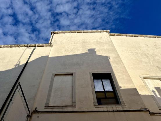 Colour photo taken looking up a high ivory coloured wall, which is in three sections, all with slightly different heights and angles, giving a jagged line of roof which runs across the frame at about a third of the way from the top. In the centre of the frame are two tall rectangular windows: the left one filled in with the same material as the rest of the wall so only the thin frame stands out in relief, and the one on the right has a white curtained top half and what looks like brown cardboard in its bottom half. A drainpipe runs horizontally below, making it look a bit like a winking robotic face. The top third of the wall is in bright glowing sun, but the shadow from the building opposite falls across the lower two thirds with sharp contours, giving the robot a shadowy face, with the shadow of a short chimney stack like a button on the top of its head. The sky above is bright blue, with some white smoky clouds on the left.