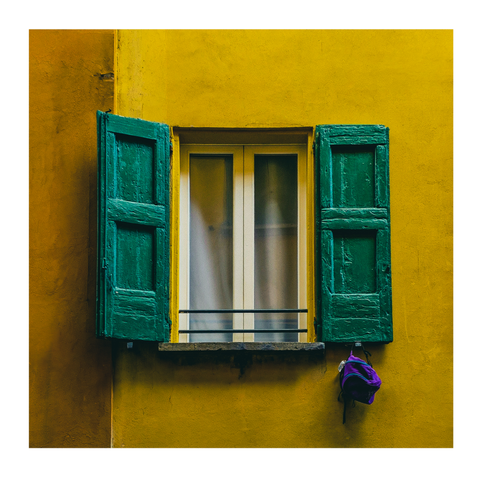 The photo is of a brightly colored yellow wall with a window in the middle of it. The window has closed glass panes with white trim and open, rustic and weathered green wooden shutters on either side. The window has cream white curtains inside. Hanging from the bottom right shutter is a small purple backpack.