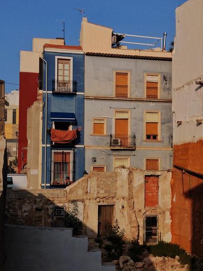 En primer plano se puede ver un solar con un muro en el que aún se ven puertas y ventanas de la antigua casa. Justo detrás hay una fachada de color azul, con ventanas y balcones amarillos. En uno de ellos hay ropa tendida.