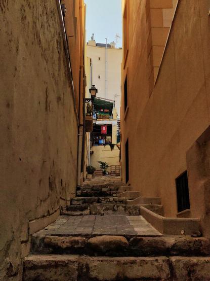 Una callejuela estrecha en el casco viejo de Alicante. Al fondo se ve una balconada con plantas y banderolas.