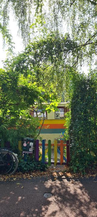 A tiny, wooden gate painted in rainbow colors between a hedge and some wild bushes and trees  The hanging branches, highlighted by the sun coming from behind, create an arch like entrance.  

Behind the gate a short, steel grate path leads down to a partly visible houseboat. The  houseboat is white, with horizontal bands of yellow, orange and blue bands running along the ship. 

In one of its windows a black and white, woodcut style poster of a  dove with the words peace now.