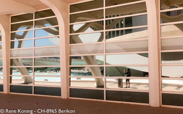 The image features a modern architectural structure with large glass windows and white, curved columns. The windows are divided into horizontal and vertical sections, creating a grid-like pattern. The reflections in the glass show the building's unique design, including white, flowing structures that resemble abstract tree branches. Outside the glass, a person is visible, standing near a trash bin, capturing a photograph. The scene includes a pool area with a bridge in the background, under a clear blue sky. The ground is paved with light-colored tiles, and the overall color palette is neutral, with white, beige, and blue tones.