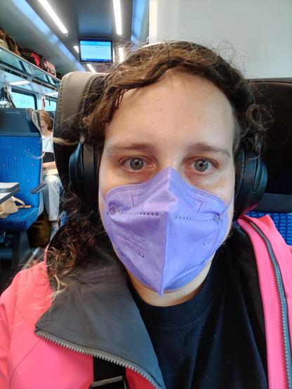 Mirah, sitting in a České dráhy train. She's wearing a black shirt, pink rain jacket, lavender mask, and headphones