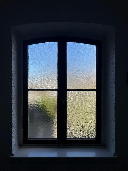 das foto zeigt ein fenster mit mattiertem, strukturiertem glas in einem dunklen raum. das fenster ist in einen bogen eingefasst und besteht aus vier einzelnen rechteckigen scheiben. durch das glas ist ein blauer himmel und ein grüner fleck von vegetation zu sehen. der kontrast zwischen dem dunklen inneren des raums und dem hellen licht von außen schafft eine ruhige und fast meditative stimmung. das bild ist einfach, aber wirkungsvoll in seiner darstellung von licht und schatten.