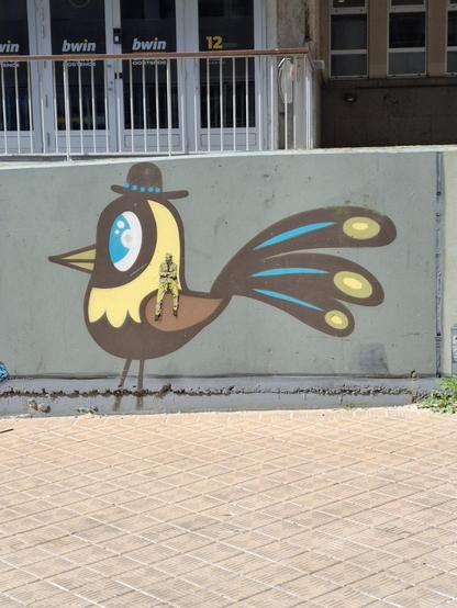 Bild eines Vogels mit gelben Kopf, schwarzem Körper, braunem Flügel  und drei Schwanzfedern in blau-schwarz-gelb. Dazu eine schwarze Melone auf dem Kopf. Auf dem Flügel sitzt ein Mann in gelben Anzug