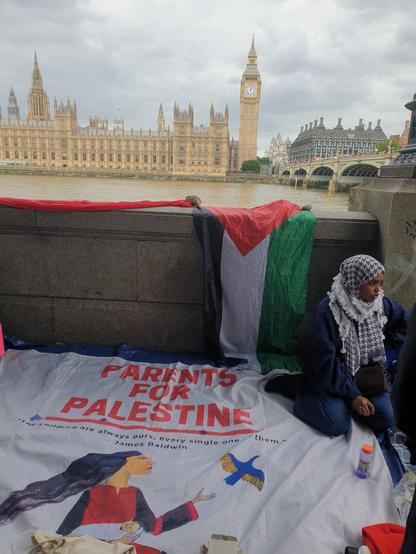Young woman in Keffiyeh sits on a banner reading 'Parents for Palestine' next to embankment in Central London. Behind her across the river are the distinctive neo-gothic Houses of Parliament.