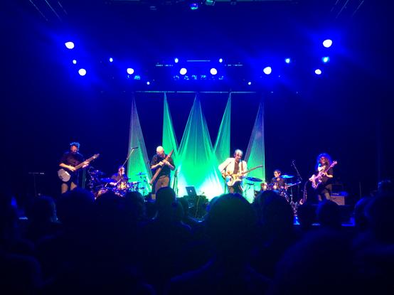 The rock bands "Stick Men" and "The Adrian Belew Power Trio" combined forces back in 2014 to go on tour as "The Crimson ProjeKCt" to play a selection of their own work interspersed - with Robert Fripp's blessing - with famous pieces from King Crimson's prodigious repertoire. The photo shows them viewed over the heads of a full house of appreciative audients. The stage is lit in a blue wash of light and five triangular pieces of fluorescent green fabric are draped across the back of the stage. The exact centre of the stage is occupied by an Apple Macbook. 

From left to right, the band are Markus Reuter, Touchguitar; Pat Mastelotto, drums; Tony Levin, Chapman Stick; Adrian Belew, guitar; Tobias Ralph, drums; and Julie Slick, bass.
