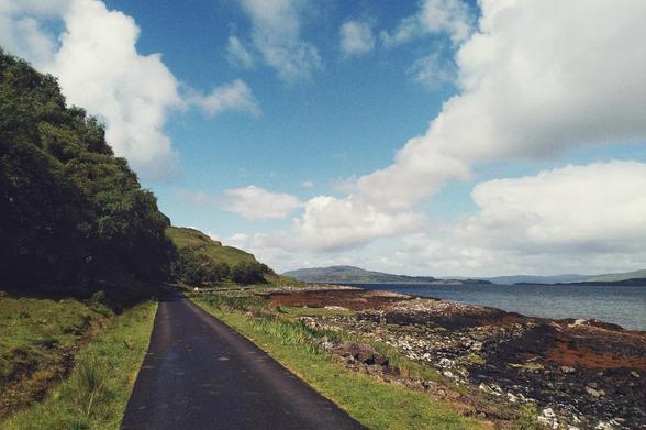 A horizontal colour photograph of a single track road bordered by grass and trees on one side, and rocks and water on the other. In the distance are some low hills. The sky is blue with big puffy white clouds.