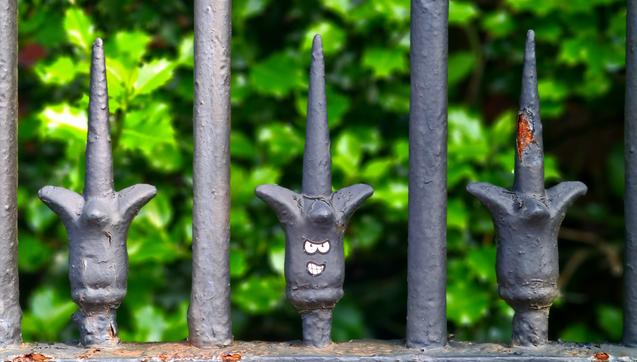 A row of three ornate spikes on a set of Victorian railings. On the middle one a pair of eyes and a mouth have been added to turn it into a head.