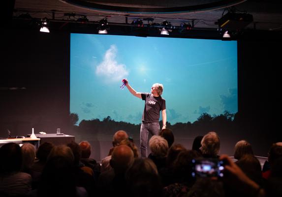 Bjorn Stevens is standing on a stage, several people in the audience are seen from behind. Stevens is spraying water into the air, from which a small cloud forms that stands out against the bright blue background.