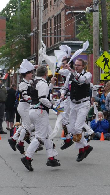 Six morris dancers with white shirts and trousers and black vests, decorated with multicolor ribbons, dancing in a street. They are in a tight group, feet off the ground and hankies in left hands raised. Behind them is an audience.