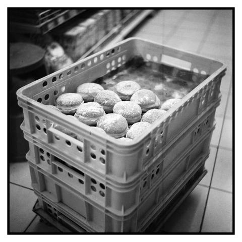 The image shows a stack of plastic crates containing doughnuts. The crates are white with circular holes along the sides for ventilation and have handles for easy carrying. The doughnuts are round, covered in a light dusting of powdered sugar, and appear to be filled, possibly with jam or cream. They are arranged neatly in rows within the top crate. The background is blurred, suggesting an indoor setting, possibly a bakery or a store, with shelves and other items faintly visible. The image is in black and white, which adds a classic and timeless feel to the scene.