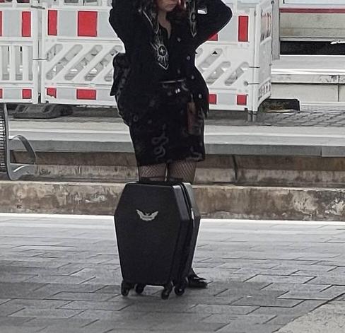 Eine junge Frau in schwarzer Kleidung an einem Bahnsteig, ihr Kopf ist absichtlich rausgeschnitten 
Vor ihr steht ein schwarzer Rollkoffer, in der typischen Sargform