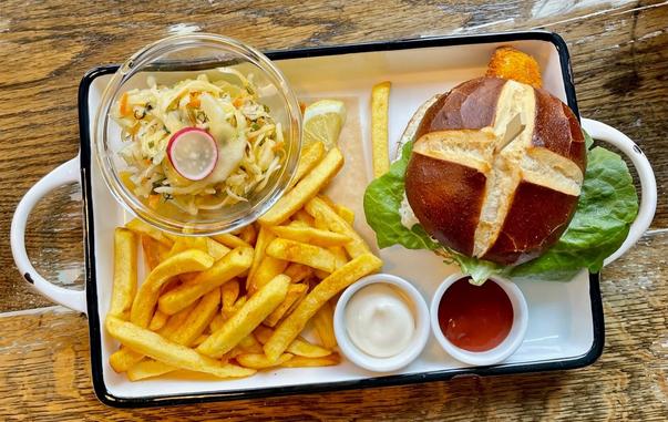 A tray holds a vegan fish sandwich on a pretzel bun with lettuce, a side of french fries, coleslaw, lemon, mayonnaise, and ketchup.