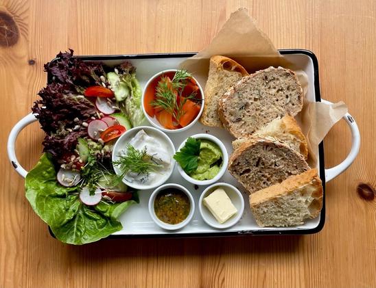 A breakfast platter with slices of bread, a green salad, various dips (including dill and pesto), pickled carrots, and butter.