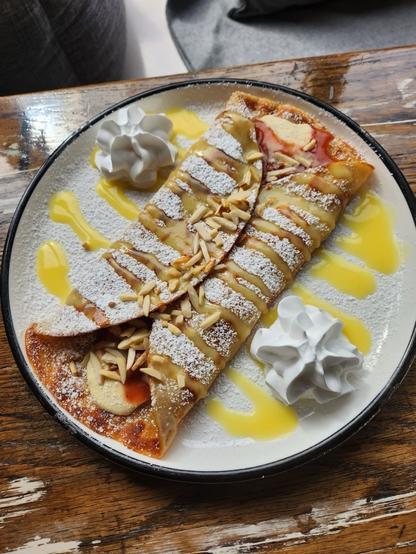 Two folded crepes dusted with powdered sugar, topped with almonds and a yellow sauce, and garnished with whipped cream.