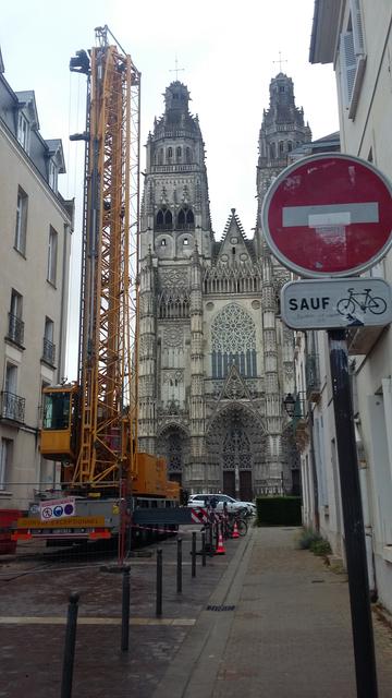engin de chantier avec une flèche élevée (segment de grue ?) devant les tours d'une cathédrale. Au premier plan, panneau de sens interdit avec "sauf cycles")