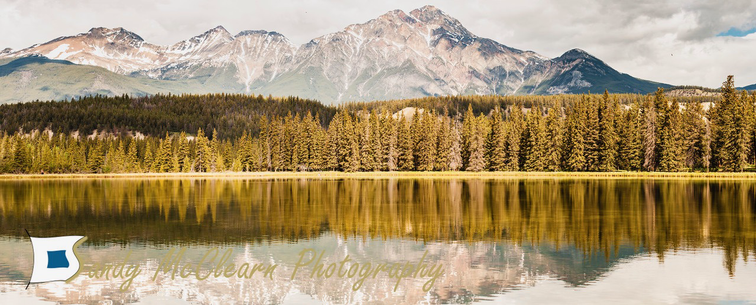 Panoramic image of trees reflecting on a calm lake against a backdrop of mountains.