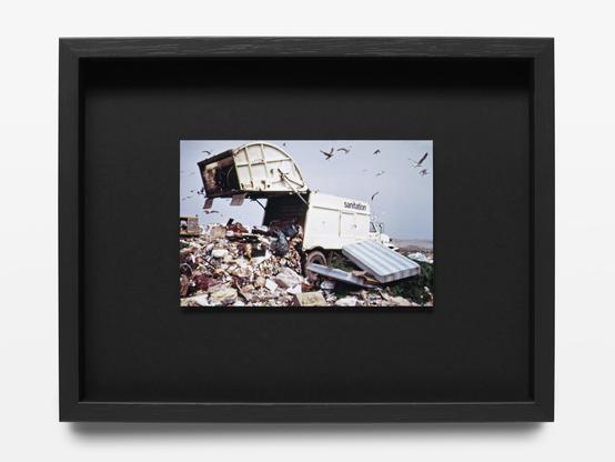 We see a coloured photograph of a sanitation truck unloading waste on a landfill. The picture was taken in 1973 in Jamaica Bay, New York.