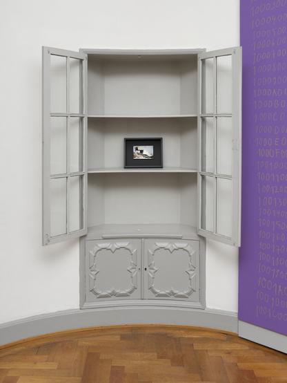 We see a grey display cabinet integrated into a wall. The wings of the cabinet are wide open. There is only a picture frame on a shelf in the otherwise empty cabinet. The frame shows a photo of a sanitation truck unloading waste at a landfill site.