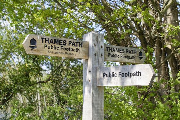 A wooden square post has three wooden pointers. Two, in opposite direction, point along the Thames path and say so. A third one is perpendicular and points to a Public Footpath. There are green trees in the background.