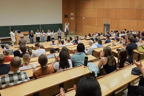 A lecture hall full of students. The students are applauding. At the front of the lecture hall you can see eight people standing in front - two are wearing lab coats, six are in plain clothes.