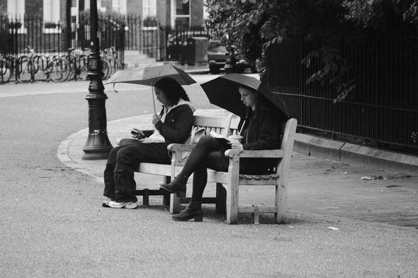 Two women sitting on a bench outside of a park in the light rain.
They both have umbrellas open, one is reading a book, the other is checking her phone.