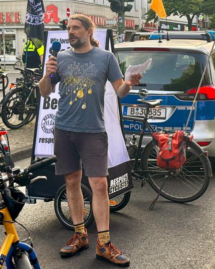 Daniel vom Netzwerk Fahrradfreundliches Neukölln hält eine Zwischenkundgebung auf der Hermannstraße mit Mikrofon und Papier vor einem Polizeiauto, umgeben von Fahrrädern und Bannern