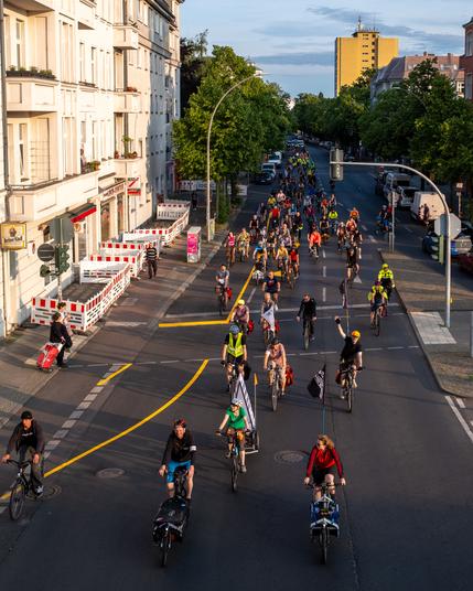 Viele Radfahrer fahren auf dem Britzer Damm in einer großen Gruppe bei Tageslicht, umgeben von Wohngebäuden und Bäumen
