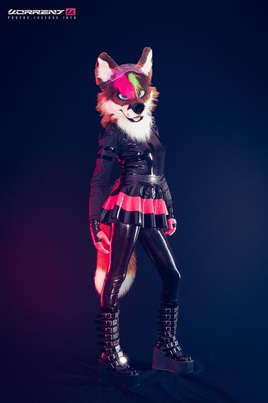 A photo of me wearing my fox fursuit head and tail, black latex catsuit under a black shrug, black and pink skirt, Demonia boots, and pink latex gloves. I'm standing and looking into the camera.