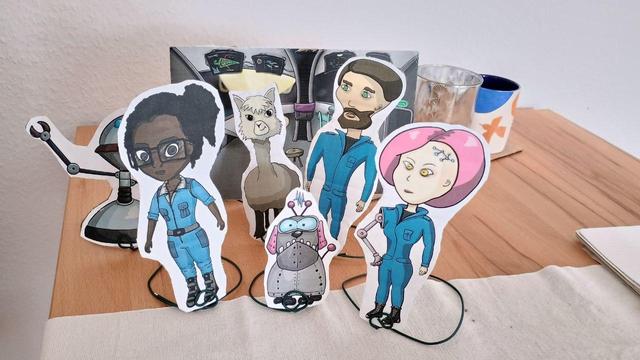 A photo of a table on which there is a glass, a cup and several cardboard stands. The cardboard stands depict three people in blue suits, an alpaca, a robot dog and a robot with long arms. A cockpit can be seen in the background.